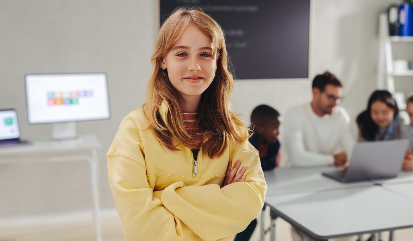 Female student attending a coding course for kids, looking the camera, while her teacher and classmates code in the background