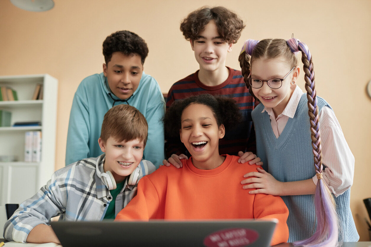 Students standing around a laptop and laughing while learning how to build app on the web without coding.